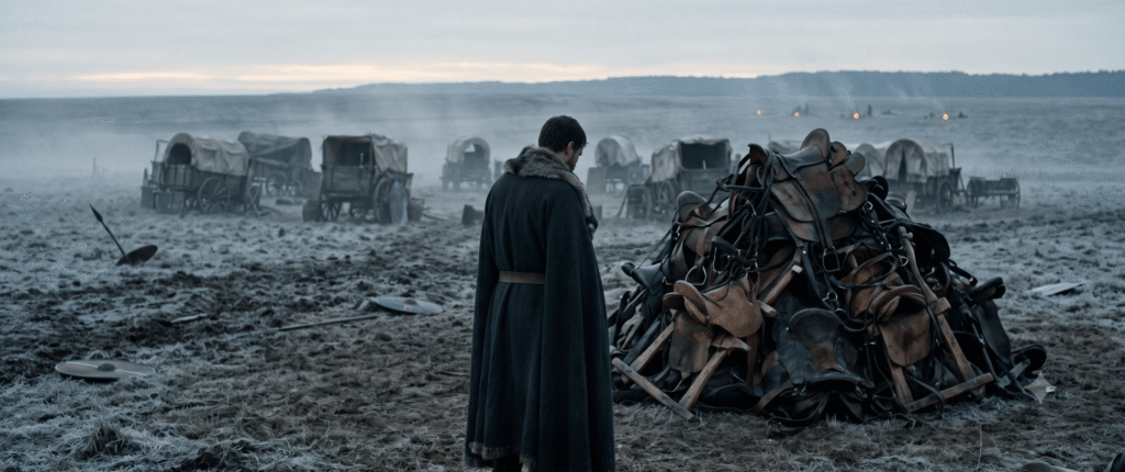 A lone figure in a dark wool cloak standing before an unlit heap of horse saddles on the cold, frosty Catalaunian Plains at dawn.